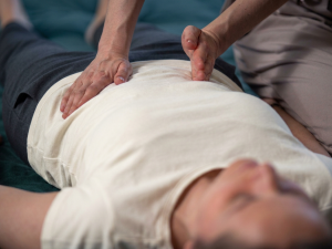 Terapeuta wykonuje masaż brzucha – terapię trzewną: jedna dłoń spoczywa na nadbrzuszu, palce drugiej delikatnie uciskają okolicę pod łukiem żebrowym, aby rozluźnić przeponę u leżącego, zrelaksowanego klienta.